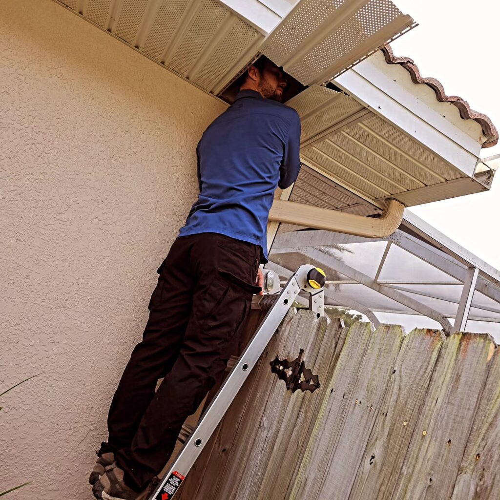 Man in a blue shirt standing on a ladder and working on a beige gutter under the roof eave outside a house.